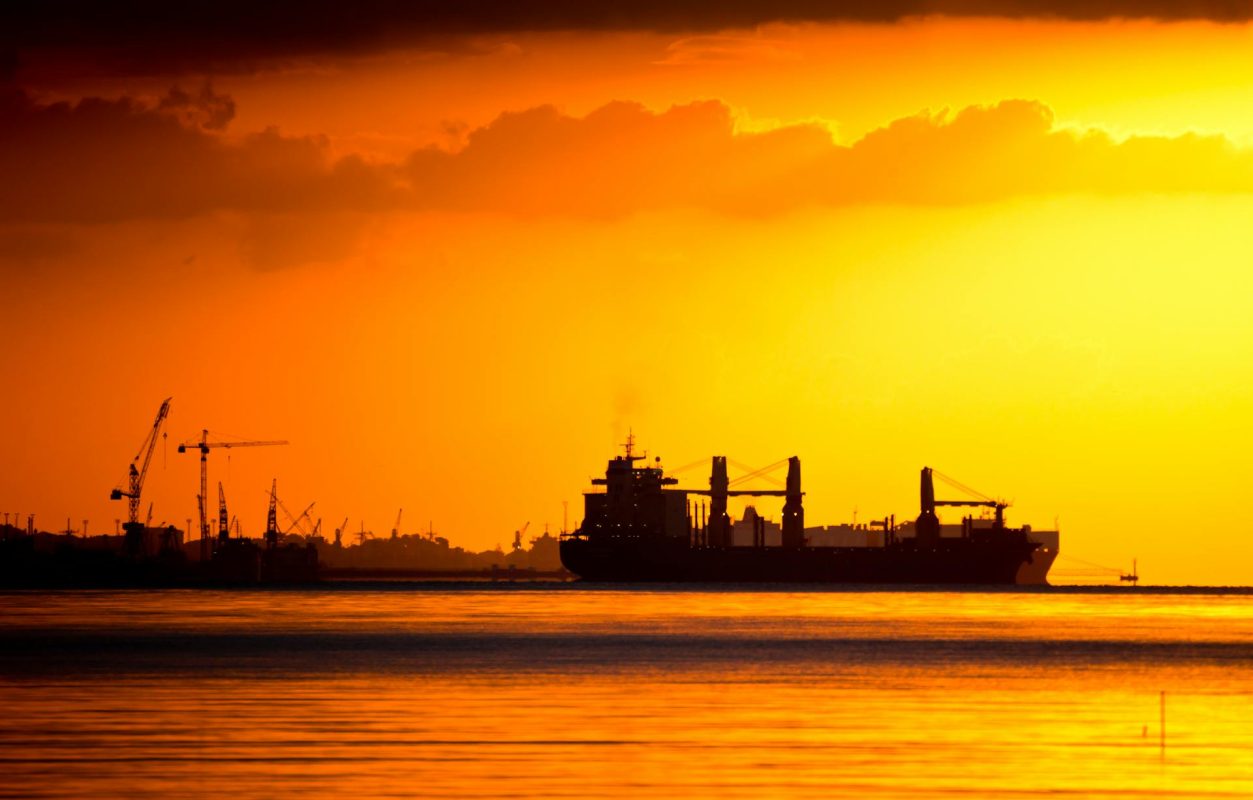 Cargo ship sailing near an industrial port with cranes at sunset.